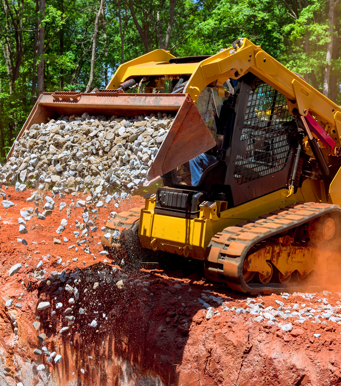 A yellow bulldozer is dumping a load of gravel into a pit on a construction site with red dirt and green trees in the background.