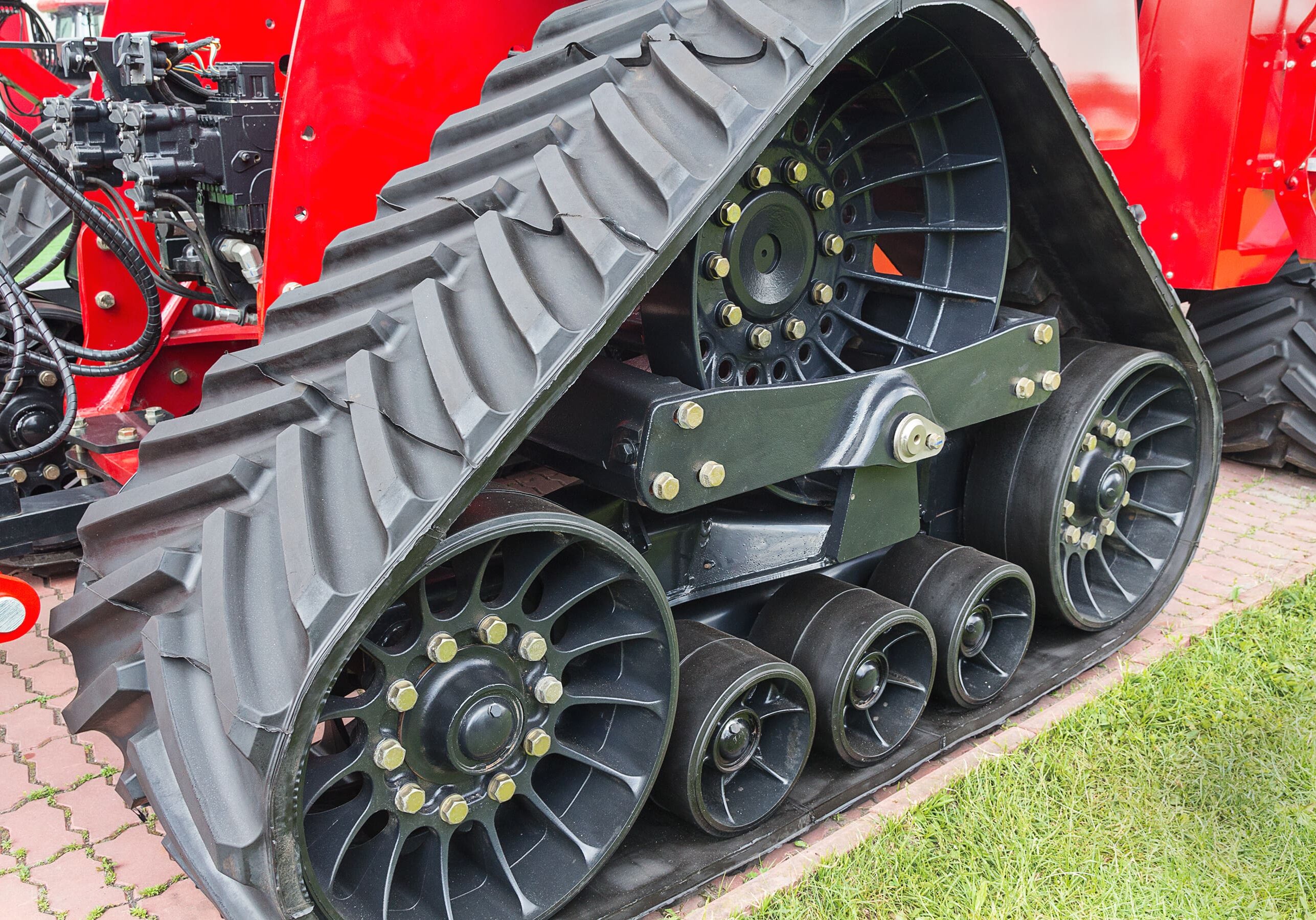 Close-up of a red tractor's rubber track system with multiple wheels and gears, on a paved and grassy surface.